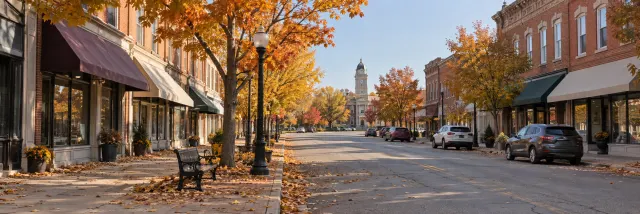 A small-town main street in October: brick storefronts, maples in orange and yellow, a bench and a lamp post.