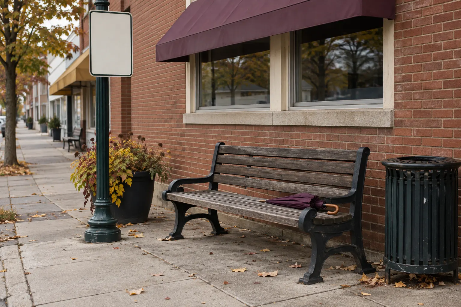 A small-town bus stop on a main street: a wooden bench with a slatted back, a blank sign on a post, a trash can, a low brick storefront with a plum-coloured awning, a folded umbrella left on the bench, leaves on the sidewalk.